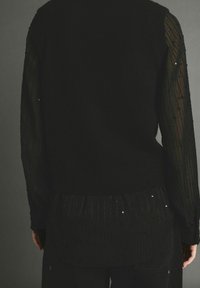 Henkilö, joka käyttää mustaa villapaitaa, jossa on läpinäkyvät, helmillä varustetut pitkät hihat ja vastaava musta toppi alla, näkyy selkäpuolelta.