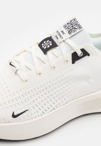 Sneaker en maille blanche avec un tissu texturé, des accents noirs et un code QR sur l'étiquette. Présente des lacets contrastants et une semelle épaisse.