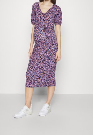 Gravid kvinde iført en tætsiddende lilla blomster midi kjole med pufærmer og hvide sneakers, stående mod en ensfarvet baggrund.