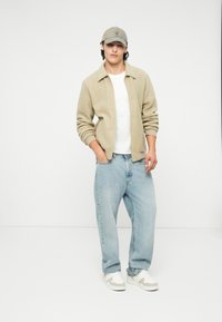 Giacca in pile beige con colletto, t-shirt bianca, jeans larghi azzurri chiari e berretto grigio. Sneakers bianche con accenti grigi completano il look.