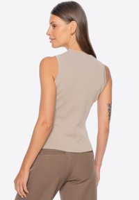 Beige žebrovaný bez rukávů top s kulatým výstřihem, přiléhavý design, nošený s hnědými kalhotami. Hladká textura, minimální detaily viditelné.