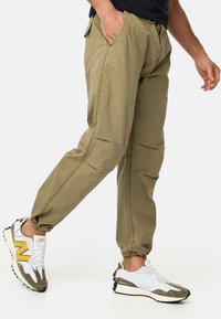 Khaki cotton jogger pants with elastic cuffs, side pockets, and stitching details, paired with white and grey sneakers featuring yellow accents.