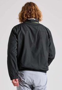 Giacca bomber nera, leggera, con polsini elasticizzati e orlo con cordino. Presenta un collo con zip e dettagli riflettenti discreti sul colletto.
