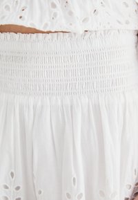 Robe en coton blanche avec un corsage smocké, des bords volantés et des découpes brodées complexes le long de l'ourlet. Texture lisse sur l'ensemble.