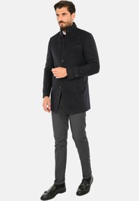 Cappotto di lana blu scuro con colletto alto, chiusura con bottoni, tasca frontale e vestibilità slim, abbinato a pantaloni grigi e scarpe nere.