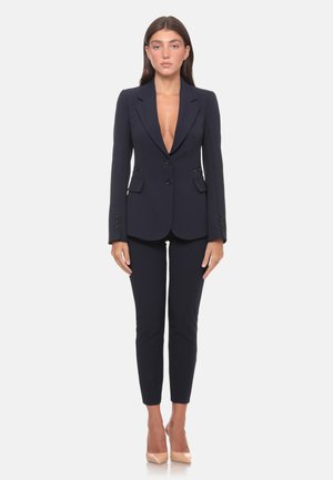 Donna in piedi che indossa un blazer nero su misura con scollatura profonda, pantaloni aderenti abbinati e tacchi alti color nude su sfondo bianco.