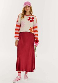 Sweter dzianinowy w kwiatowe wzory w kolorze kremowym, różowym i czerwonym, połączony z ciemnoczerwonym satynowym spódnicą i różowymi butami sportowymi. Różowa czapka i kwiaty w ręku.
