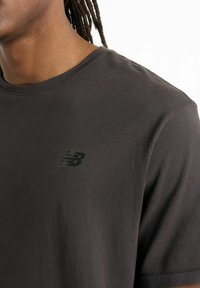 Black cotton T-shirt with a ribbed texture. Features a small black New Balance logo on the chest. Classic crew neckline and short sleeves.