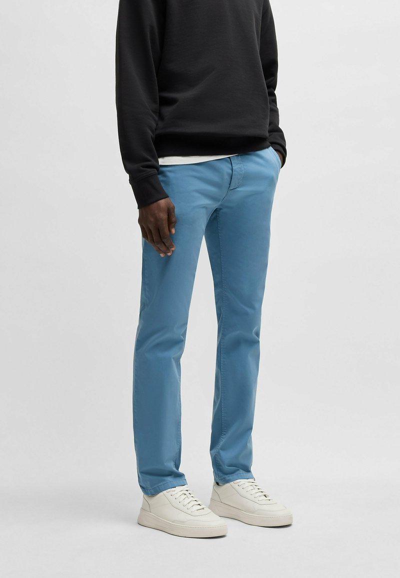 Chinos bleu clair en coton, avec une coupe slim et un léger éclat, portés avec des baskets blanches et un sweatshirt noir.
