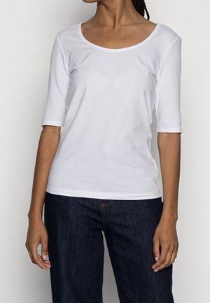 Persoon die een witte top met halflange mouwen en een ronde hals draagt, en een donkerblauwe spijkerbroek, staand tegen een eenvoudige lichte achtergrond.