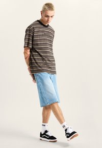 Camisa de manga corta a rayas en marrón y negro, combinada con unos pantalones cortos de mezclilla azul claro. El calzado consiste en unas zapatillas negras y blancas con suelas de goma.