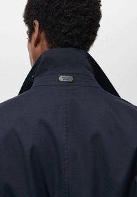 Giacca impermeabile blu navy con colletto alto, dotata di etichetta metallica nera sul retro, texture liscia e vestibilità tailored.