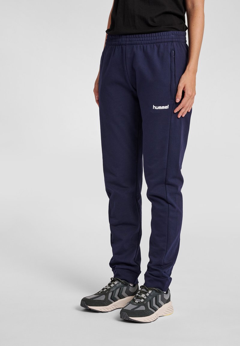 Marina blå träningsoveraller i mjukt tyg, med elastisk midja, sidoficka med dragkedja och vitt logo på vänster lår.