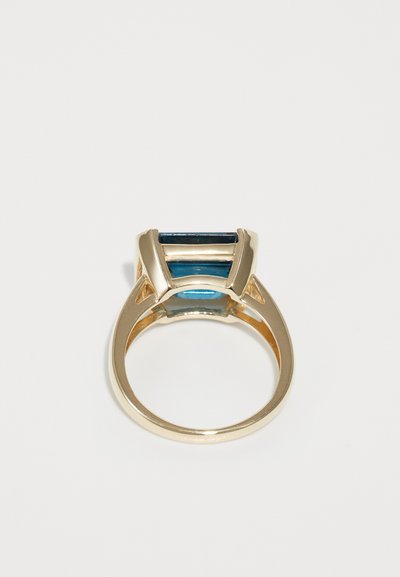Anillo de oro con un centro de piedra azul rectangular, flanqueado por dos piedras azules más pequeñas, superficie pulida y diseño moderno.