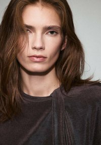 Detailný záber modelky s dlhými, vlnitými hnedými vlasmi. Má na sebe tmavý, sametový top s unikátnym detailom na pleci a prirodzený make-up.