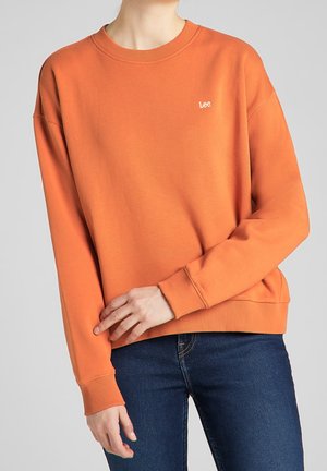 Persona con sudadera Lee naranja con puños y dobladillo acanalados, combinada con vaqueros azul oscuro, sobre un fondo gris liso.