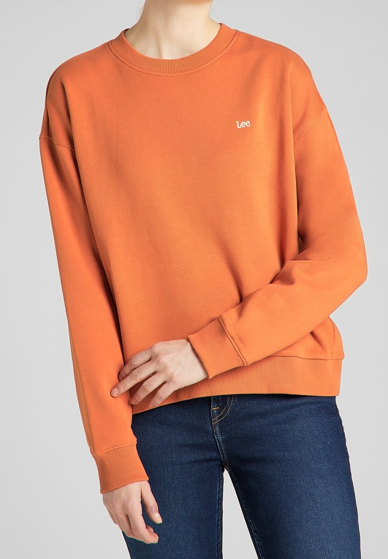 Personne portant un sweat-shirt Lee orange avec poignets et ourlet côtelés, associé à un jean bleu foncé, sur un fond gris uni.
