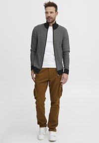 Sudadera de punto gris con cremallera y acentos negros, combinada con pantalones cargo marrones con bolsillos laterales y zapatillas blancas. Diseño minimalista.