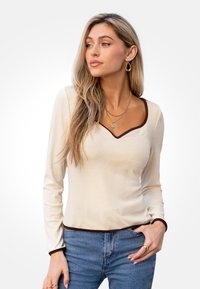 Top a maniche lunghe, a coste, di colore crema con scollo a V marrone e polsini. Abbinato a jeans blu a vita alta.