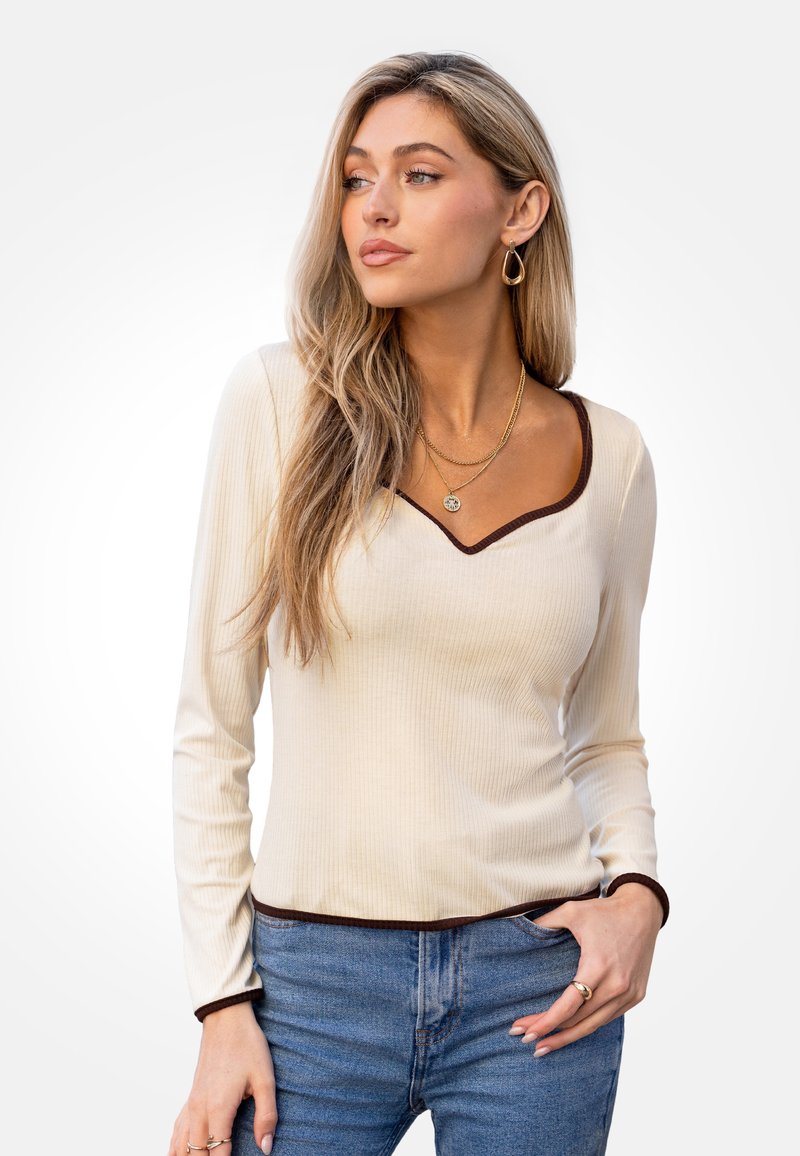Top a maniche lunghe, a coste, di colore crema con scollo a V marrone e polsini. Abbinato a jeans blu a vita alta.