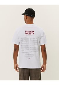 White cotton t-shirt featuring printed text on the back in burgundy. The front is plain. The wearer is also wearing a black cap.