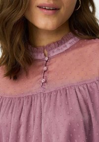 Blouse rose pâle en tissu texturé à pois, col à volants et trois boutons décoratifs à l'avant. Tissu doux et léger.