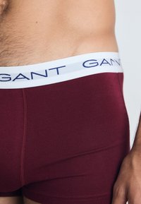 Boxer ajustados burdeos con una cinturilla elástica blanca que presenta la inscripción "GANT" en azul. Tejido suave y ceñido, con costuras planas.