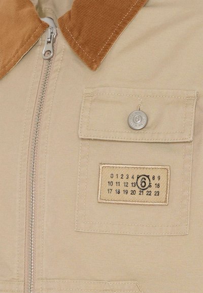 Giacca beige con colletto in velluto a coste, zip argentata e tasca frontale con etichetta cucita decorata con un motivo a calendario. Tessuto dalla texture liscia.