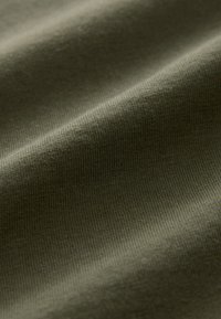 Close-up van zachte, olijfgroene stof met fijne gebreide textuur en subtiele plooien die zachte schaduwen creëren.