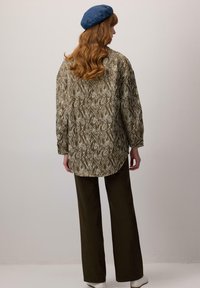 Camicia oversize a fantasia verde con maniche lunghe, abbinata a pantaloni a gamba larga verde scuro e un berretto blu, vista da dietro.