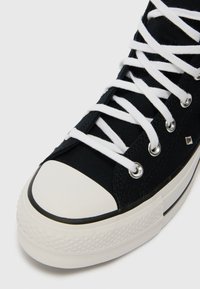 Sneaker înalt din pânză neagră, cu vârf din cauciuc alb, talpă groasă albă și șireturi albe trecute prin ochiuri metalice.