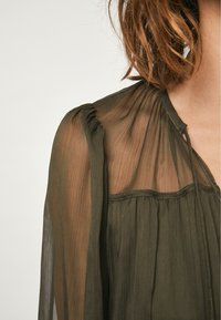 Donker olijfgroen sheer blouse met delicate plooien, een zachte ronde halslijn en subtiele gathers bij de schouders. Lichtgewicht, vloeiende stof.