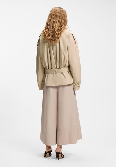 Femme aux cheveux blonds bouclés portant une veste beige ceinturée, un pantalon fluide beige clair et des sandales à talons noires, vue de dos.