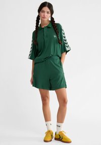 Zielona, prążkowana koszulka polo z krótkim rękawem, z logo i wzorem chevron. Połączona z dopasowanymi zielonymi szortami i żółtymi trampkami.