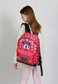 Mochila rosa con lunares blancos, gráfico de Minnie Mouse, bolsillo frontal con cremallera y base negra, y parches decorativos en rojo y amarillo.