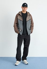 Větrovka v šedé a hnědé barvě, černé tričko, tmavé džíny a tenisky. Výrazná kapuce, detaily na zipu a logo na bundě.