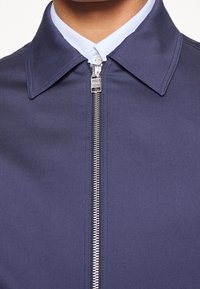 Marinblå jacka med en strukturerad krage, med en hel frontdragkedja och textil med struktur. Synlig sömnad förbättrar hållbarheten.