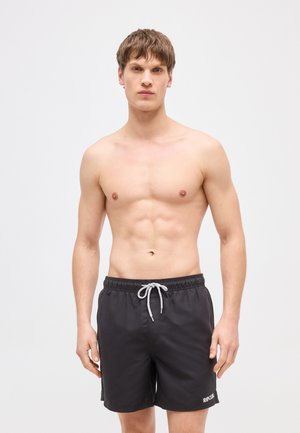 Jeune homme aux cheveux bruns courts portant un short de bain noir avec un cordon blanc, debout devant un fond clair uni.