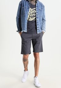 Jeansowa kurtka, czarna koszulka z grafiką, ciemne szorty i białe sneakersy. Charakteryzuje się wytatuowanymi nogami i luźnym krojem.