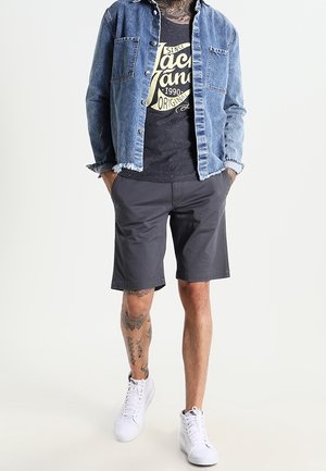Spijkerjack, zwart graphic t-shirt, donkere korte broek en witte sneakers. Heeft getatoeëerde benen en een casual pasvorm.