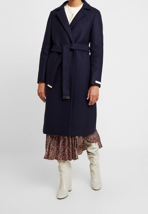 Femme portant un manteau en laine bleu marine foncé ceinturé, une jupe à motifs et des bottes blanches montantes, debout devant un fond uni.