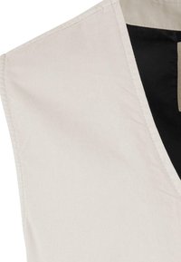 Gros plan d’un gilet en tissu beige avec des coutures visibles et une doublure intérieure noire au niveau du col.