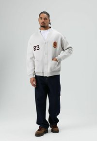 Witte katoenen cardigan met knoopsluiting, geborduurd embleem en nummer 23. Gecombineerd met donkere spijkerbroek en bruine veterlaarzen.