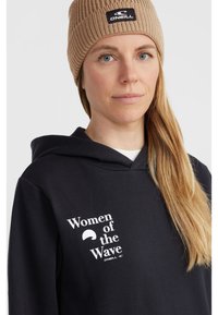 Black hoodie featuring white text "Women of the Wave" and logo, worn with a tan knit beanie. Hair is long and straight.