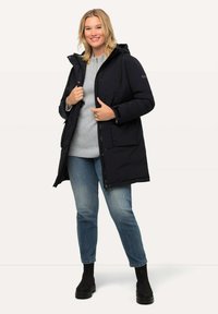 Chaqueta tipo abrigo navy con capucha, cierre frontal y bolsillos laterales, combinada con jeans azul claros y botas negras, contra un fondo neutro.