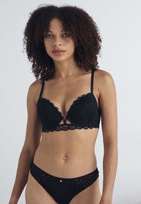 Bralette in pizzo nero con bordi ondulati, caratterizzato da un dettaglio a chiave sul davanti e spalline regolabili. Abbinato a mutandine trasparenti coordinate.