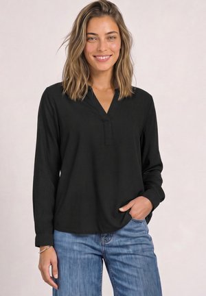 Donna sorridente con capelli alla spalla indossa top nero a maniche lunghe con scollo a V e jeans blu, posa con una mano in tasca.