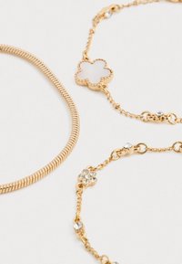 Kolme kultasävyistä rannetta: joustava käärmeenergia, kukkakoru teksturoidulla pinnalla sekä yksi, jossa on strassikoristeita ketjussa.