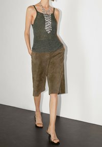 Un débardeur en maille vert olive associé à des culottes en suède marron. La tenue est complétée par un long collier en argent et des talons ouverts noirs.