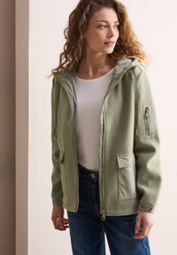 Giacca verde oliva con cappuccio, chiusura a zip e due tasche frontali. Presenta un tessuto di colore verde più chiaro in contrasto sul cappuccio e sulle maniche.